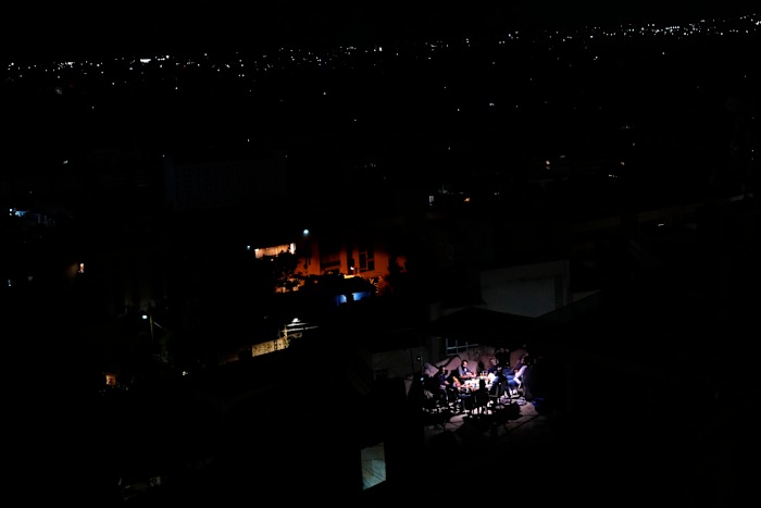 Bewoners zitten op het dak van een gebouw en dineren terwijl de buitenwijk Dahiyeh, op de achtergrond, in duisternis blijft na Israëlische luchtaanvallen, Libanon, vrijdag 11 oktober 2024. (AP Photo/Hassan Ammar)